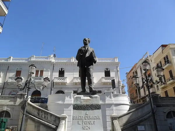 Bronze statue of Biagio Camagna in Piazza Camagna, framed by Liberty-style buildings and a semicircular staircase in Reggio Calabria.